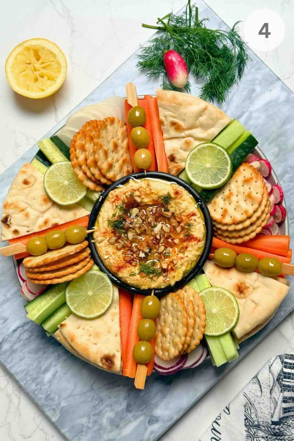 Arranging fresh raw vegetables, olives, biscuits, and a bowl of dip on a platter.