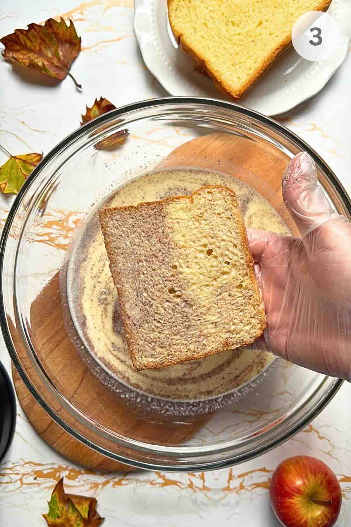 Thick brioche slice being dipped into custard in a shallow dish.