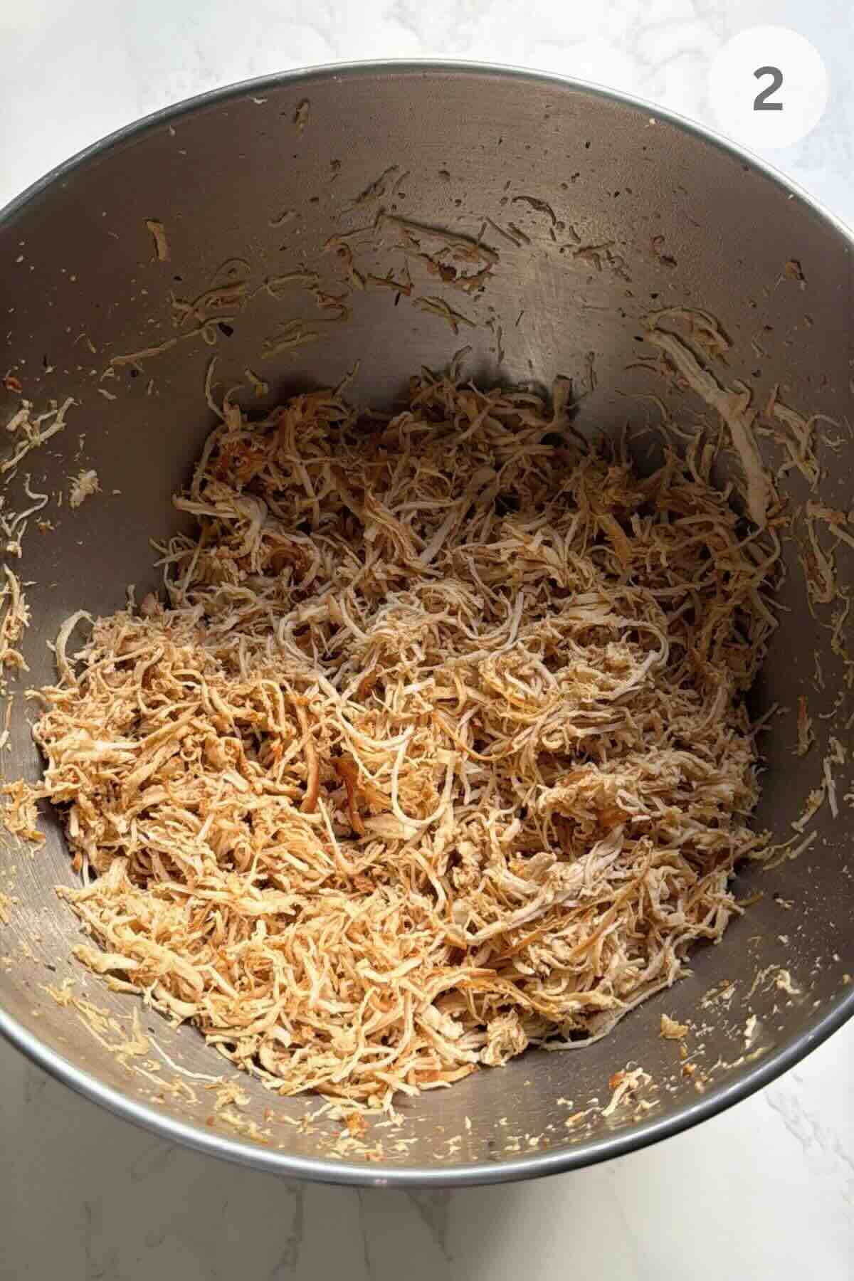 Shredding cooked chicken breast with a hand mixer in a mixing bowl.